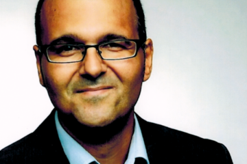 Amin Salim. Grinsender Mann mit dunkler eckiger Brille, einem hellblauen Hemd und schwarzem Sakko. Zuständig für Coaching, Fortbildungen, Moderation & Supervision.