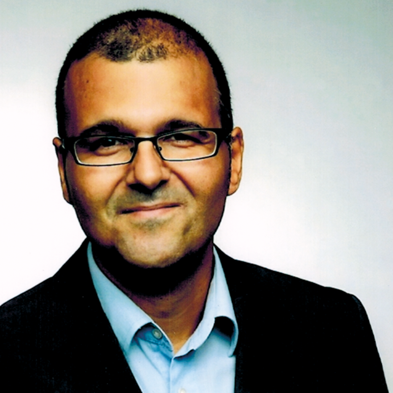 Amin Salim. Grinsender Mann mit dunkler eckiger Brille, einem hellblauen Hemd und schwarzem Sakko. Zuständig für Coaching, Fortbildungen, Moderation & Supervision.