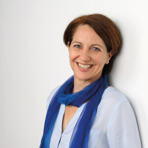 Christiane Muth. Frau mit braunen schulterlangen Haaren, einem blauen Seidenschal und hellblauer Bluse. Zuständig für Coaching, Fortbildungen & Supervision.