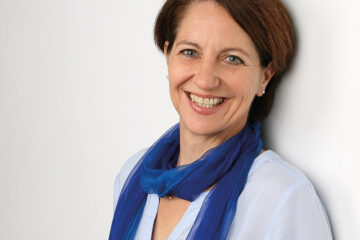 Christiane Muth. Frau mit braunen schulterlangen Haaren, einem blauen Seidenschal und hellblauer Bluse. Zuständig für Coaching, Fortbildungen & Supervision.