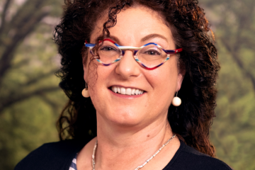 Jeanette Piram. Eine Frau mit lockigen braunen Haaren, weißen Kugelohringen und einer stylishen Brille. Zuständig für Beratung, Coaching, Fortbildungen & Supervision.